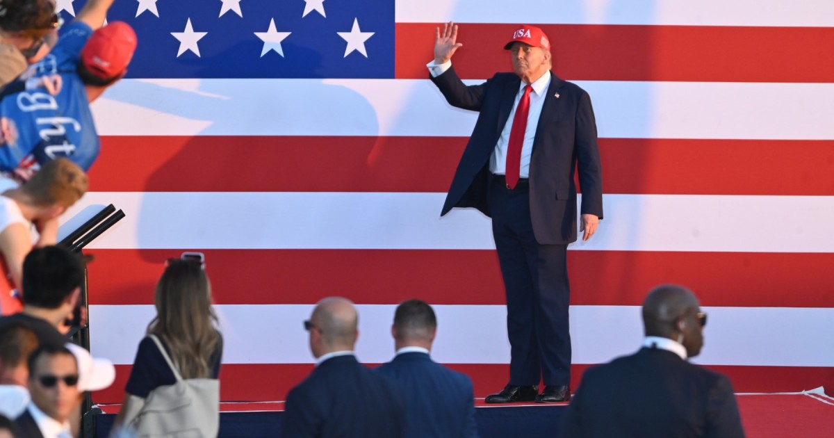 Donald Trump pe o scenă, îmbrăcat cu un costum bleumarin, cămașă albă și cracată roșie, pe cap are o șapcă roșie cu „MAGA”, face cu mâna către mulțime. În spatele lui, un panou cu steagul Americii.