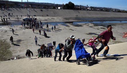 Migranți se grăbesc să traverseze albia secată a râului Tijuana în încercarea de a escalada gardul spre Statele Unite. Foto: Northfoto