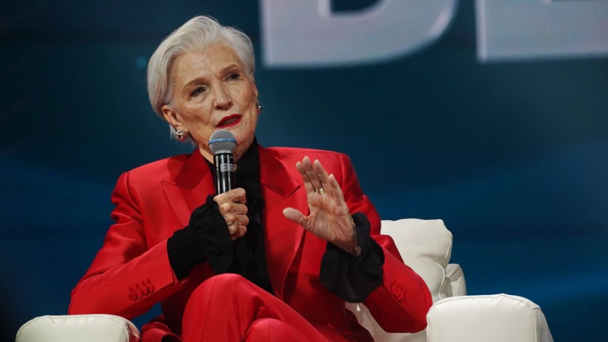 Maye Musk, mama lui Elon Musk, la o conferința de presă, îmbrăcată într-un costum roșu din două piese, cu o cămașă neagră