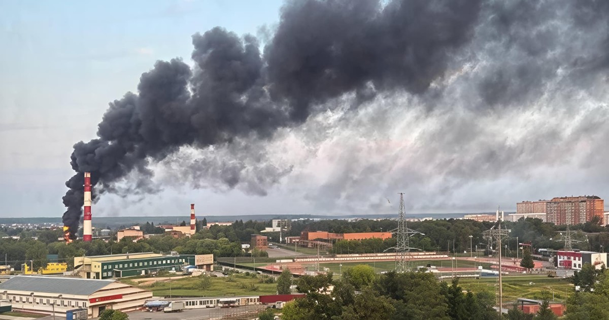 o vedere panoramică dintr-un punct înalt asupra unei zone cu profil mixt, industrial și rezidențial. În partea stângă a cadrului, exact la baza unui coș de fum industrial foarte înalt, are loc un incendiu puternic cu flăcări pronunțate. Din acest focar se înalță o coloană masivă și extrem de densă de fum, care se extinde pe orizontală și acoperă o mare parte din jumătatea superioară a compoziției. În prim-plan și în zona centrală a peisajului se observă hale cu acoperișuri plate, porțiuni extinse de vegetație cu copaci, precum și un complex sportiv cu un teren de dimensiuni mari înconjurat de o pistă de alergare. În partea dreaptă, în depărtare, se profilează un ansamblu de clădiri rezidențiale masive, cu mai multe etaje. Peisajul este completat de prezența unor stâlpi înalți pentru rețele electrice de înaltă tensiune și a unui al doilea coș de fum, de dimensiuni mai reduse, situat în planul îndepărtat.