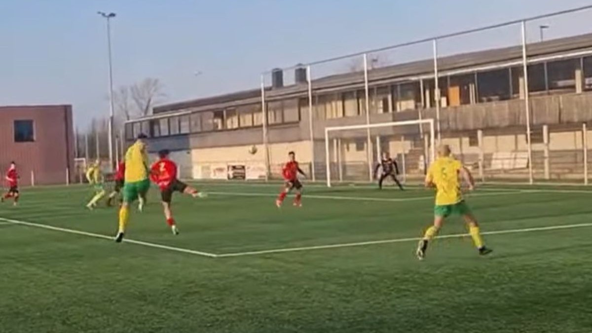 o fază dintr-un meci de fotbal desfășurat pe un teren cu gazon artificial verde, delimitat de linii albe clare. În centrul acțiunii, mai mulți jucători sunt dispersați pe teren: unii poartă echipamente galbene cu șorturi verzi, în timp ce adversarii lor sunt îmbrăcați în tricouri și șorturi roșii. Mingea este vizibilă în aer, în apropierea careului de pedeapsă, unde un jucător în roșu pare să fi executat o lovitură. În fundal, se află poarta de fotbal cu plasă albă, în fața căreia este poziționat portarul, iar mai departe se întinde o clădire lungă cu un etaj, prevăzută cu numeroase ferestre mari de sticlă și un acoperiș plat