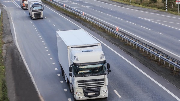 Un camion alb de mare tonaj circulând pe banda unei autostrăzi cu mai multe benzi, urmat la distanță de alte autovehicule comerciale.   