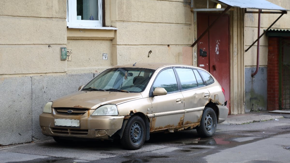 O mașină foarte veche, are caroseria ruginită și este parcată pe marginea unei străzi