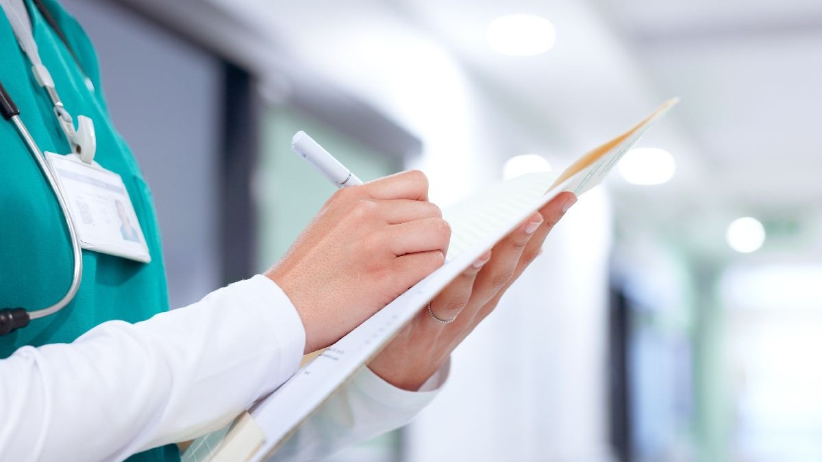 Imaginea prezintă un cadru în prim-plan cu mâinile unui cadru medical care notează informații pe o fișă de observație medicală, având un stetoscop la gât și ecusonul la vedere.