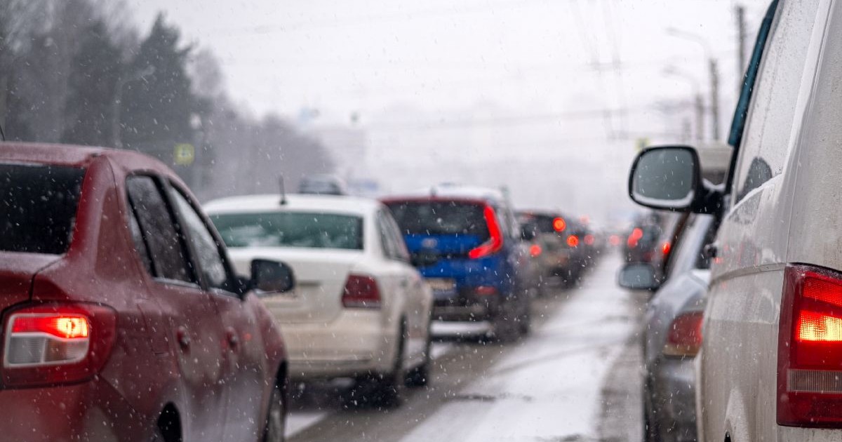 O stradă acoperită de zăpadă, pe care se vede o coloană de vehicule, inclusiv mașini roșii și albe, care se deplasează încet pe drumul alb.
