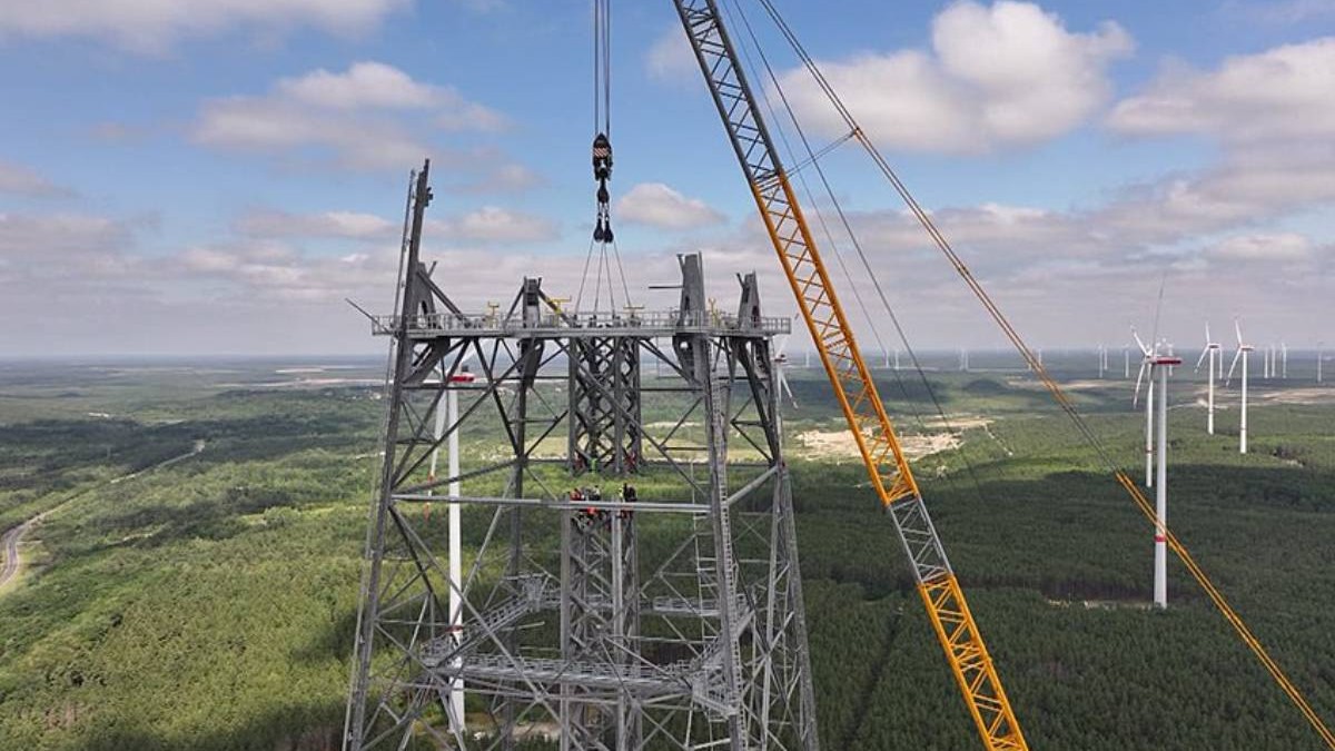 Imagine aeriană realizată în timpul construcției unei turbine eoliene uriașe în Germania. În prim-plan se vede structura metalică masivă a turnului, aflată încă în montaj, iar o macara foarte înaltă ridică elemente grele deasupra acesteia. În fundal se întind păduri și câmpuri, printre care sunt vizibile mai multe turbine eoliene convenționale, mult mai mici.
