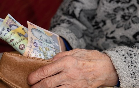 O bătrână cu pulover pe ea scoate dintr-un portofel maro, pe care îl ține cu o mână, câteva bancnote românești