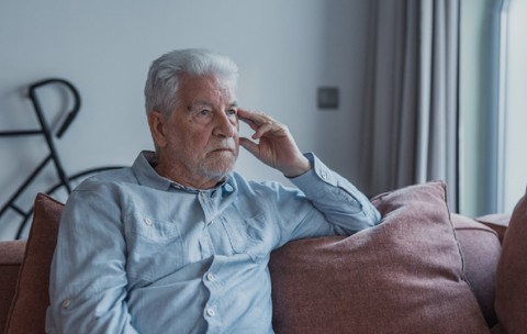 Bărbatul în vârstă stă singur pe o canapea privind afară cu o expresie tristă