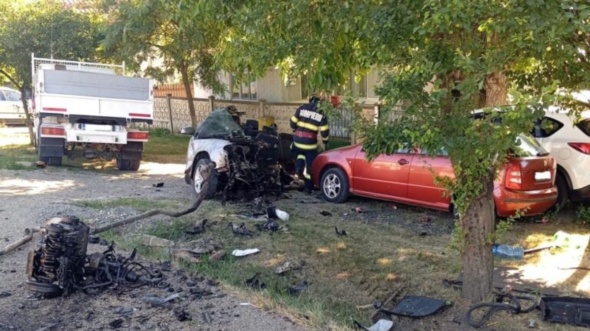 un autoturism alb, complet distrus în partea din față, cu motorul și alte componente aruncate la câțiva metri distanță de caroserie. Lângă autoturism se află un pompier îmbrăcat în uniformă roșie și pe marginea drumului, sunt parcate alte două mașini, una roșie și una albă, care par să fi fost avariate ușor.