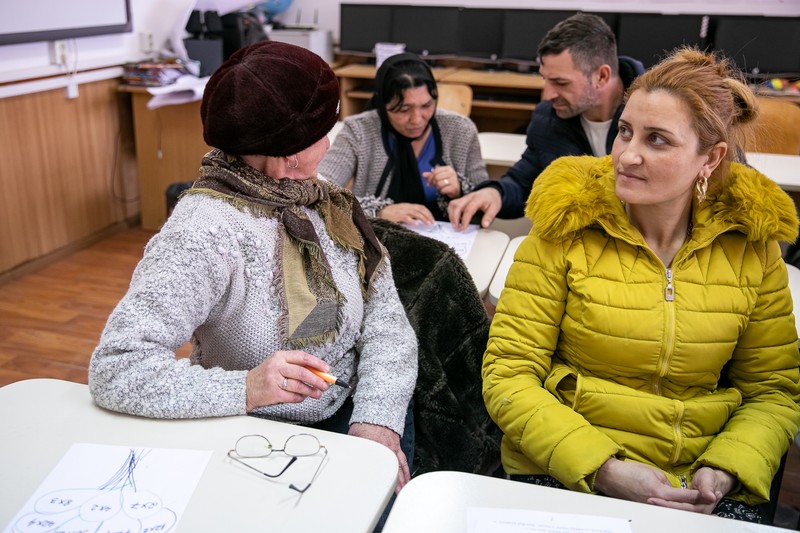 Eugenia, 38 de ani, în bancă. În spate, un bărbat o ajută pe colega lui să rezolve un exercițiu