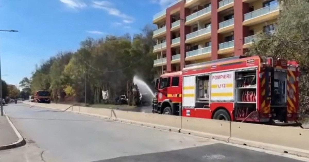 Imaginea surprinde o scenă de urgență în care o mașină de pompieri roșie din România, cu inscripția "Pompierii 112", pulverizează apă asupra unui focar de incendiu. Acțiunea se desfășoară în fața unei clădiri rezidențiale cu mai multe etaje, lângă o alee mărginită de copaci, probabil în cartierul Pipera din București, așa cum sugerează numele fișierului.