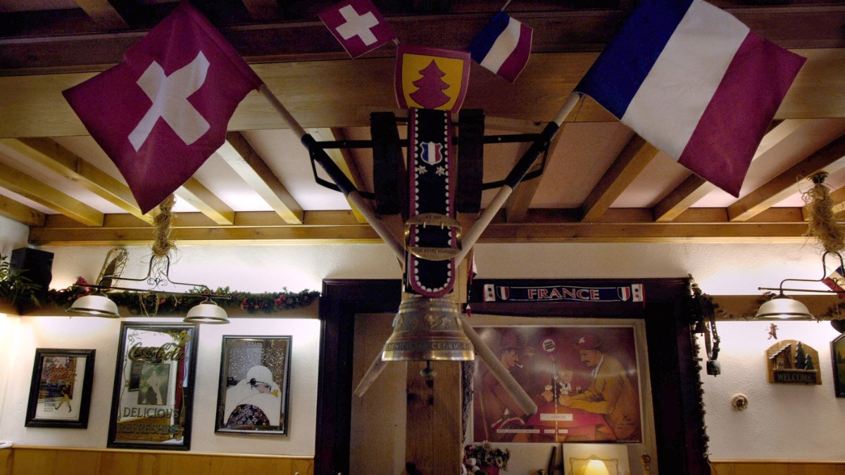 Imaginea surprinde interiorul unui restaurant rustic, decorat cu steagurile Franței și Elveției, sugerând clar poziționarea sa chiar pe granița dintre cele două țări. Elementele tradiționale din lemn și obiectele decorative subliniază atmosfera caldă, istorică și caracterul unic al locului.