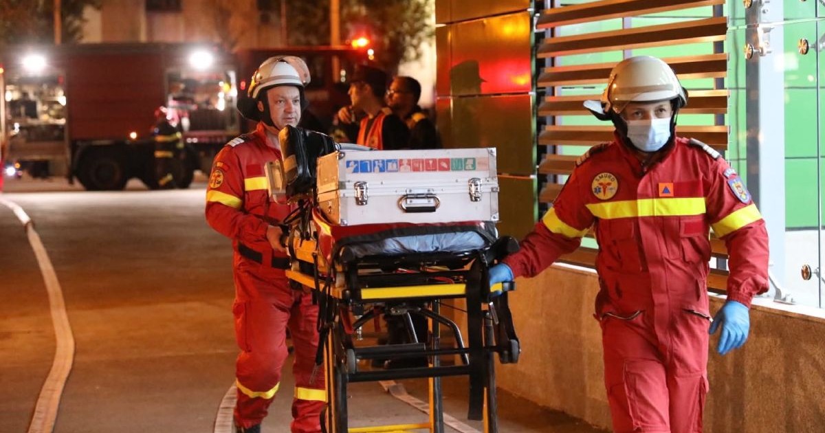 In imagine doi salvatori SMURD înbrăcați în combinezoane rosii transporta echipament pe o targă, seara, în cadrul Exercițiului SEISM 2025 Convex 3