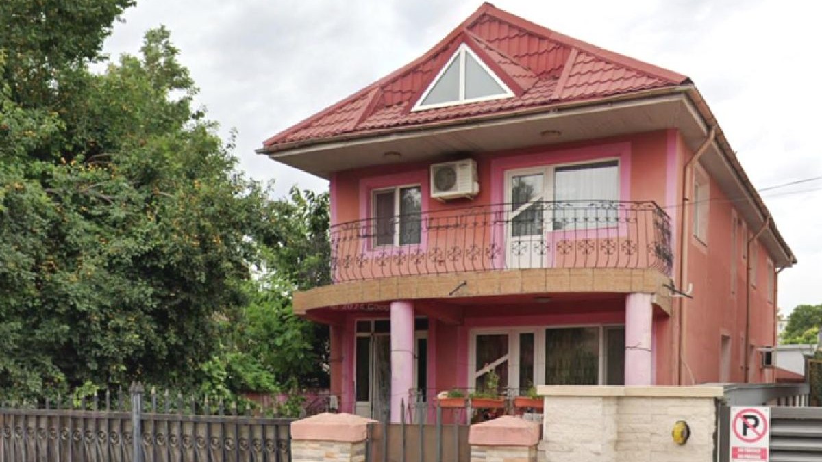 Fotografie cu o casă roz cu etaj, acoperiș roșu. Vila este înconjurată de copaci verzi și un gard din cărămidă.