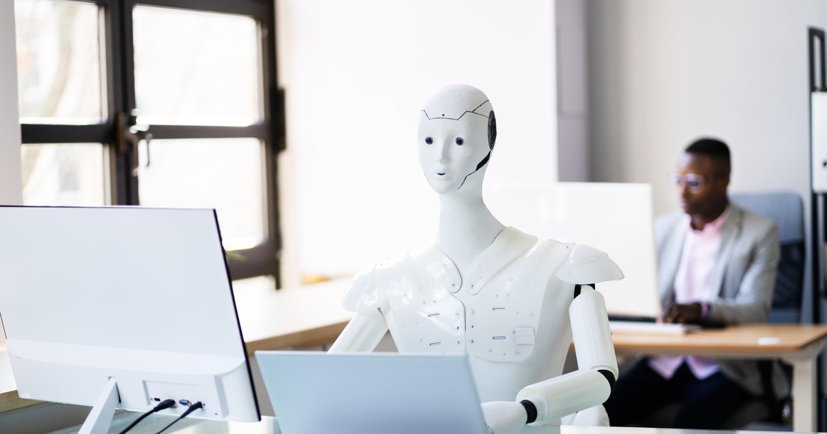 Imagine cu un robot alb stând la birou în fața unui laptop și a unui monitor. În spate se află un alt om care privește într-un monitor