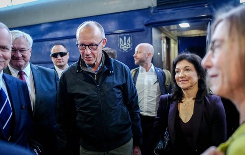Friedrich Merz purtând o jachetă închisă la culoare și ochelari, încadrat de oficiali și însoțitori lângă un vagon de tren ucrainean.