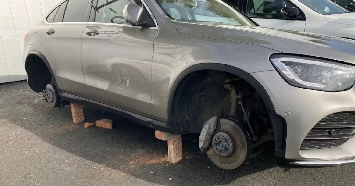 Un Mercedes-Benz argintiu, cu roțile lipsă, este sprijinit pe cărămizi într-o parcare, cu un alt vehicul parțial vizibil în lateral.