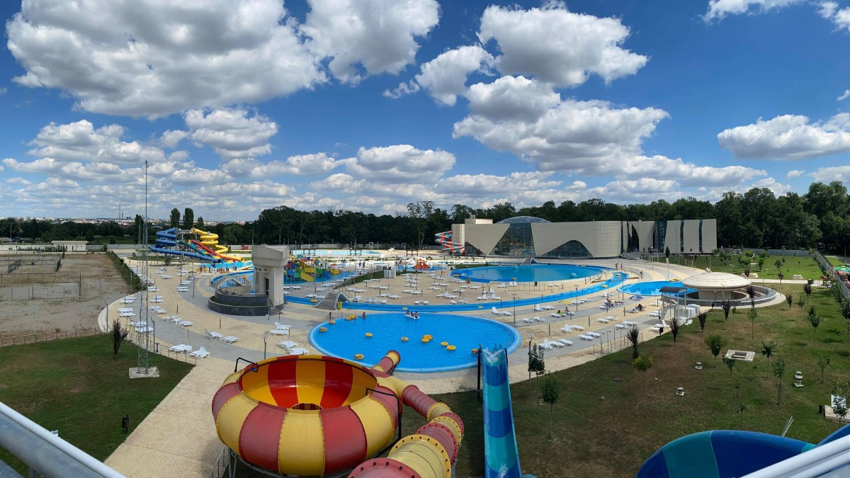 Imaginea surprinde un parc acvatic modern, cu piscine spațioase, tobogane colorate și numeroase șezlonguri, într-o zi însorită cu cer senin și nori pufoși. Complexul este înconjurat de spații verzi și pădure, oferind un cadru ideal pentru relaxare și distracție în sezonul estival.