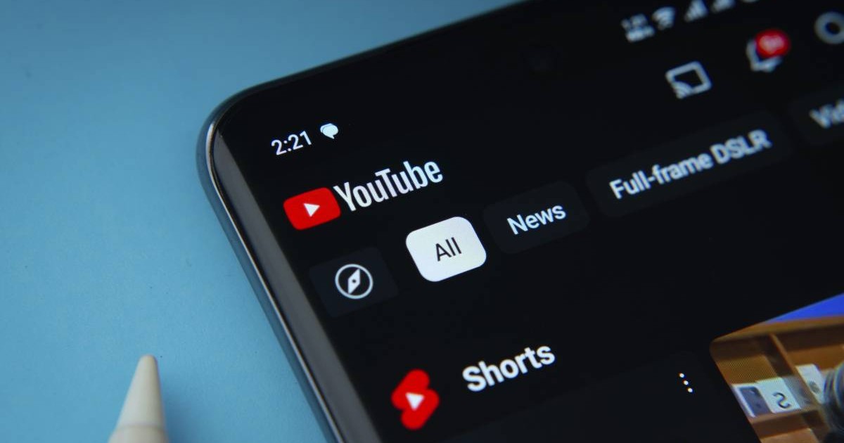 Aplicația YouTube, deschiusă pe ecranul unui smartphone așezat pe un fundal albastru deschis.