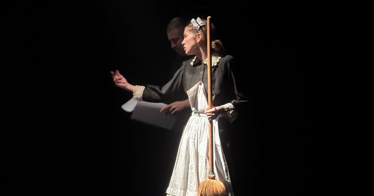 Imaginea surprinde o actriță pe scenă, îmbrăcată în costum de cameristă, ținând o mătură și gesticulând expresiv într-o lumină dramatică. Decorul întunecat și jocul de lumini pun accent pe interpretarea sa, sugerând o scenă intensă și teatrală.