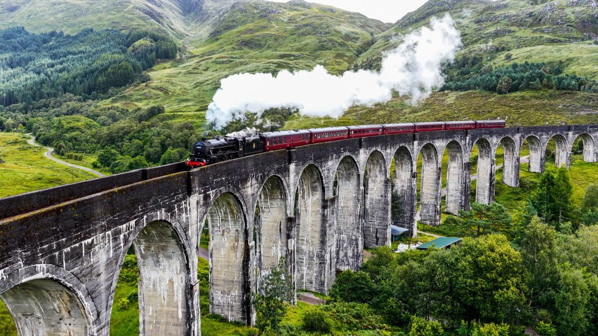 Vedere aeriană a viaductului Glenfinnan, cu un tren cu aburi care traversează arcadele istorice din piatră din Highlands-ul scoțian; punct de referință turistic emblematic, înconjurat de dealuri verzi și peisajul văii; concept de turism și patrimoniu