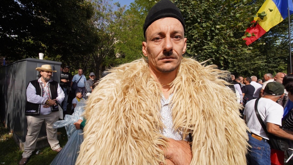 Un bărbat purtând o căciulă neagră și un cojoc din blană de oaie, privind fix spre cameră în mijlocul altor participanți la un protest în aer liber, unde se observă și un steag al României.