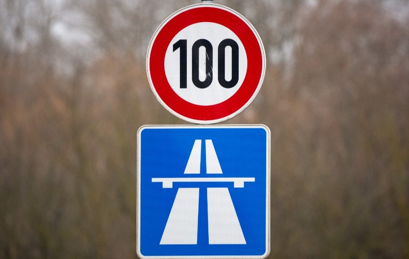 Indicator rutier de limitare a vitezei la 100 km/h, amplasat deasupra unui panou albastru care semnalizează intrarea pe o autostradă.   