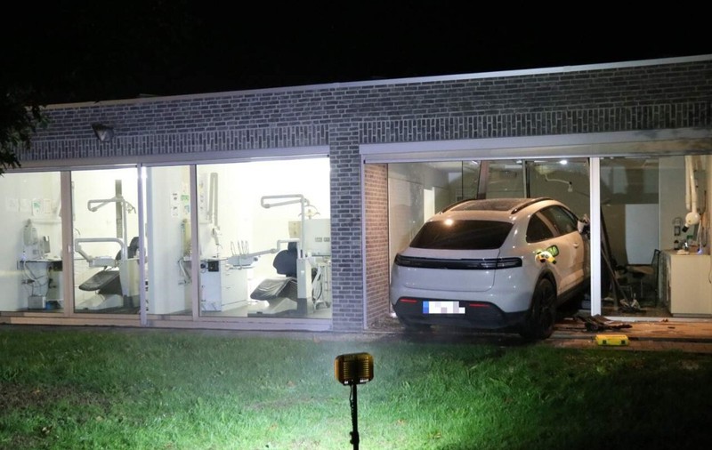 Un SUV Porsche alb care a intrat prin peretele de sticlă al unei clădiri moderne în care funcționează un cabinet stomatologic, pe timp de noapte.