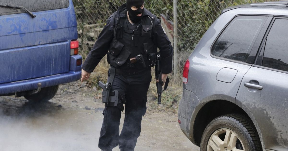 Un polițist mascat, îmbrăcat în negru, cu o armă în mână, vesta antiglonț și pistoale la el, este în misiune și trece pe stradă printre două mașini