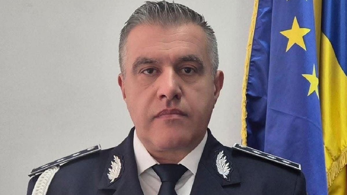 Fotografie a unui bărbat de vârstă mijlocie, cu părul scurt și cărunt, îmbrăcat în uniformă închisă la culoare, cu epoleți argintii și cravată neagră, stând în fața unui steag.
