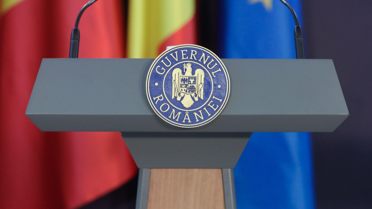 Un pupitru oficial de conferințe ce poartă stema și inscripția „GUVERNUL ROMÂNIEI”, dotat cu două microfoane, având pe fundal steagul României și cel al Uniunii Europene.