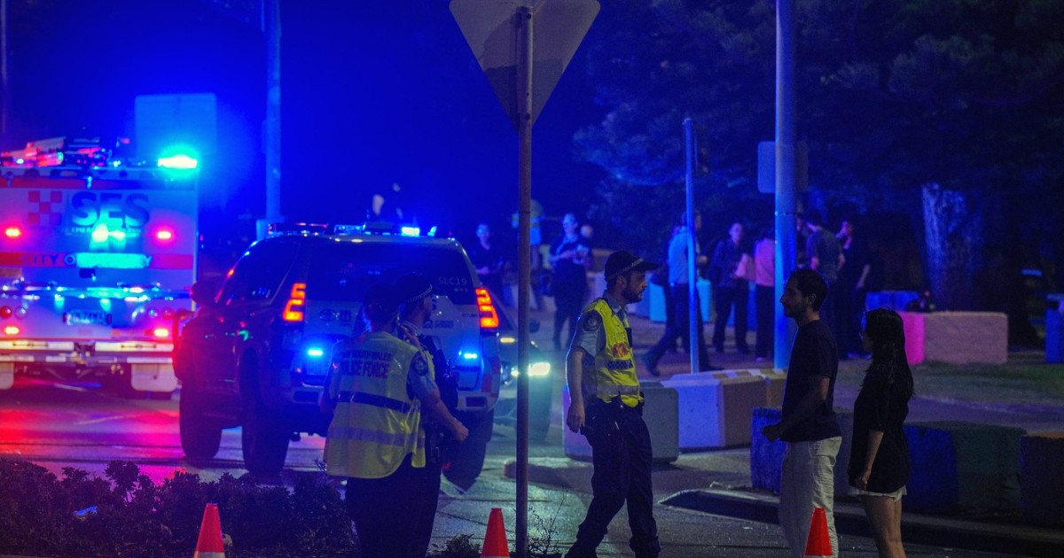 Imaginea surprinde o intervenție a forțelor de ordine pe timp de noapte, într-o zonă urbană din Australia. Polițiști și echipaje de urgență, cu girofarurile aprinse, au delimitat perimetrul cu conuri reflectorizante, în timp ce discută cu civili aflați la fața locului, sugerând o situație de securitate sau un incident grav în desfășurare.