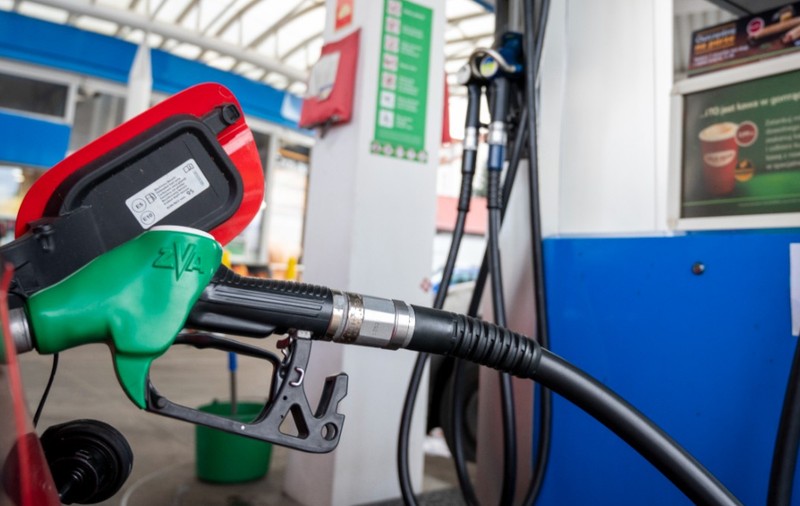 Imaginea prezintă un pistol verde de pompă de carburant introducător în rezervorul deschis al unei mașini roșii la o stație de alimentare.   