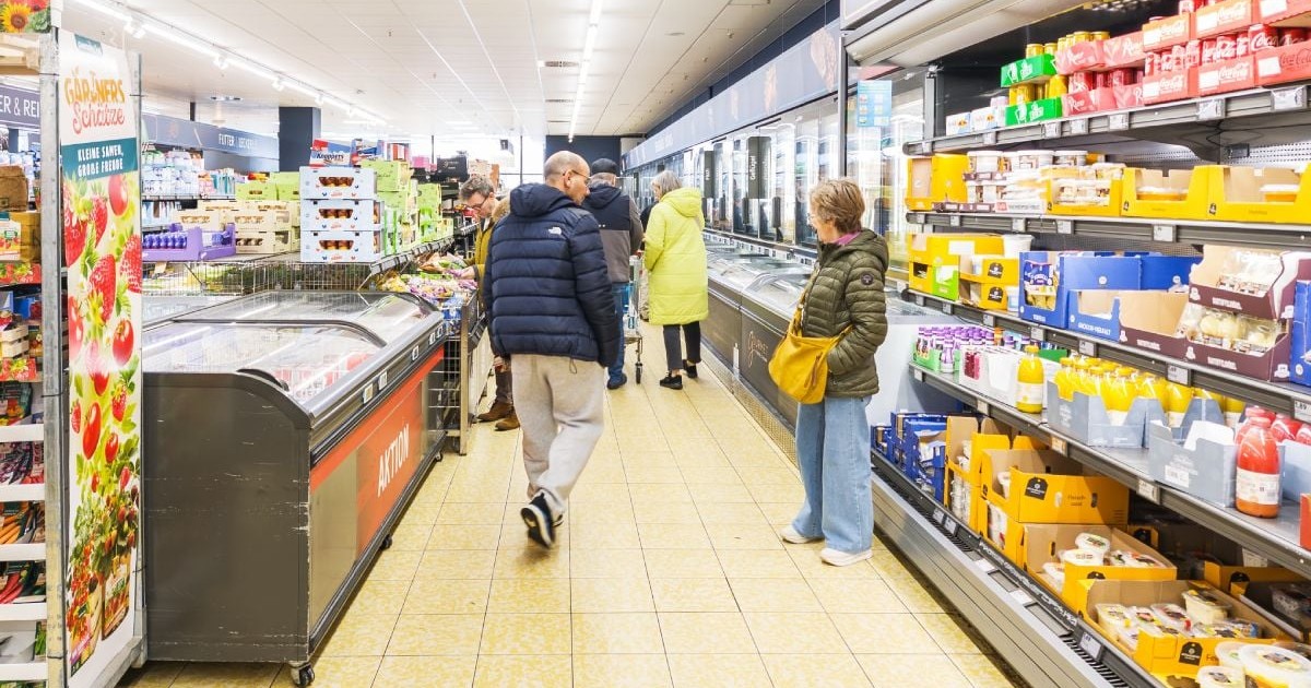 Imaginea prezintă mai mulți clienți care fac cumpărături pe culoarul unui supermarket, examinând produsele din vitrinele frigorifice și de pe rafturile pline cu alimente și băuturi.