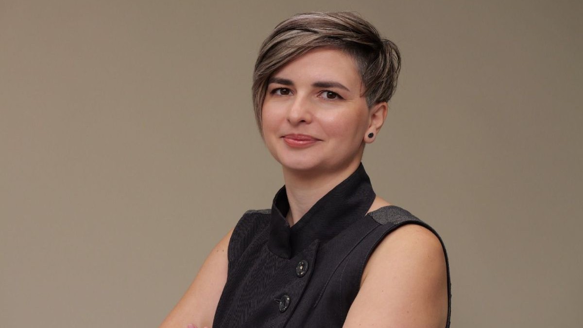 Roxana Cristolovean, în prim plan, îmbrăcată cu o bluză neagră, fără mâneci, iar pe un fundal un perete de culoare crem închis
