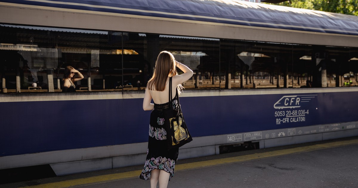 o femeie văzută din spate, stând în picioare pe peronul pavat al unei gări, orientată cu fața către un vagon de tren de călători oprit. Ea poartă o rochie lungă de vară, decupată la spate și cu un model floral în partea inferioară, purtând sandale și având o sacoșă din pânză cu un imprimeu grafic pe umăr. Femeia are o mână ridicată la nivelul frunții, iar în ferestrele mari și reflectorizante ale vagonului i se observă clar reflexia chipului și a gestului, alături de reflectarea stâlpilor și a structurii peronului de vizavi. Pe partea exterioară a vagonului, sub geamuri, este inscripționat logoul „CFR” însoțit de mai multe serii numerice și texte tehnice, în timp ce pe asfaltul peronului, paralel cu trenul, este trasată o linie continuă de demarcație.