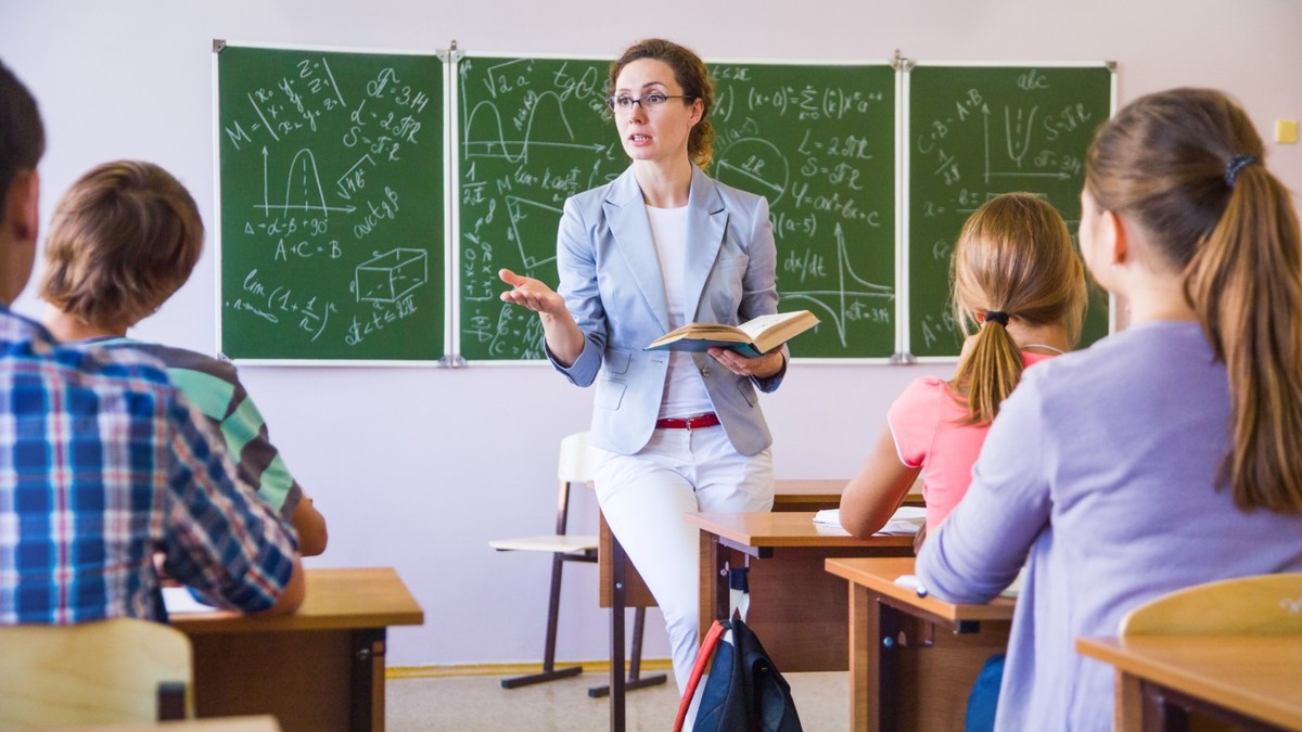 Elevi în sala de clasă, fotografiați cu spatele, iar în fața lor este profesoara care explică ceva și în spate se vede tabla verde pe care este scrie cu cretă albă la matematică