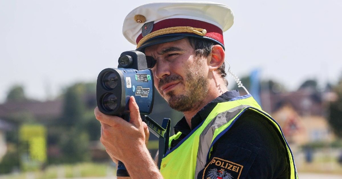 Un bărbat cu barbă, îmbrăcat în uniformă de polițist și cu o șapcă albă cu cozoroc ține un parat radar la ochi