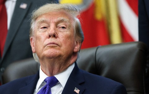 Donald Trump privind în sus, îmbrăcat într-un costum închis la culoare, cămașă albă, cravată albastră cu model și un pin cu drapelul Statelor Unite la rever.