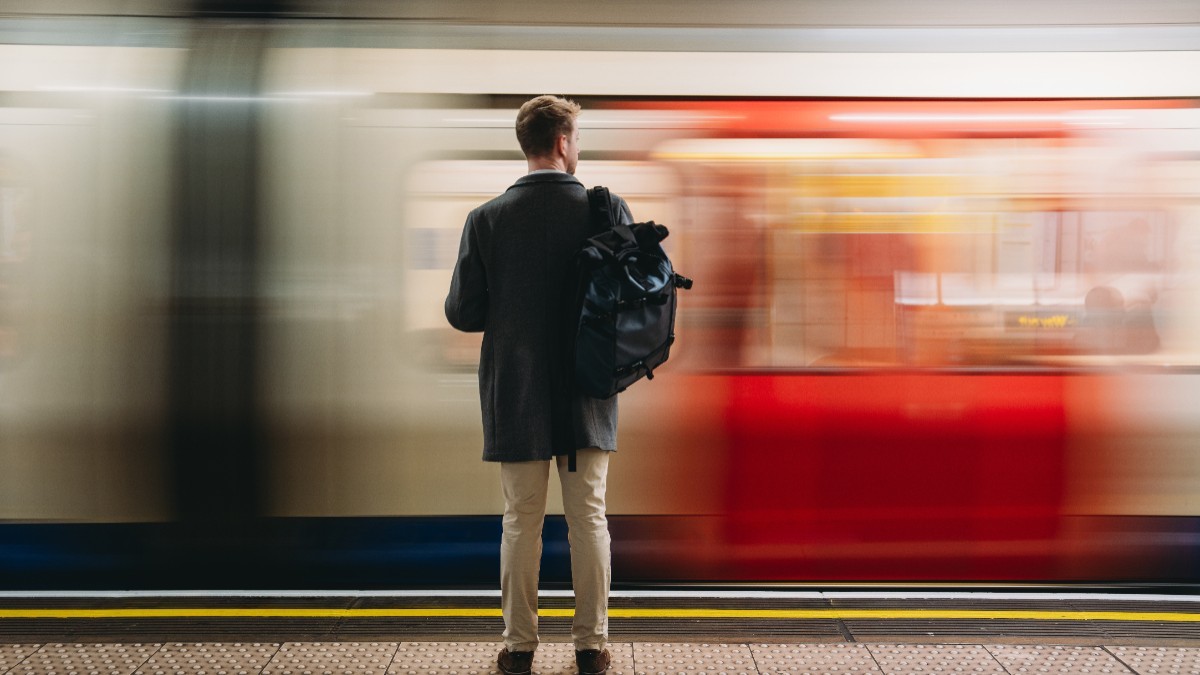 Un bărbat cu rucsac în spate, văzut din spate pe peronul unei stații de metrou, în timp ce prin fața lui trece un tren în viteză.