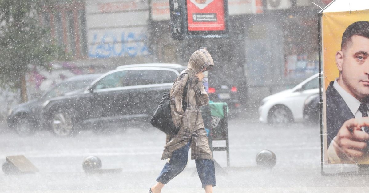 O femeie surprinsă de vremea rea în București merge pe stradă, se protejează de ploaie cu gluga și trece pe lângă un afiș electoral. Foto: Libertatea/Dumitru Angelescu