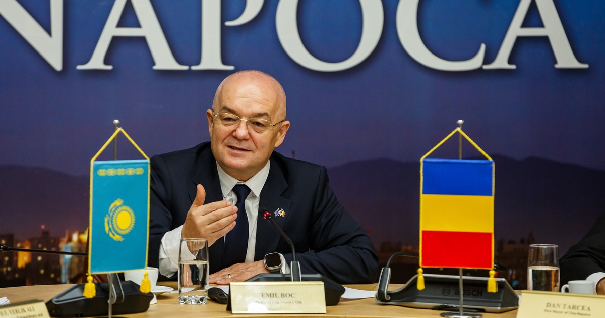 Primarul din Cluj, Emil Boc, stă la o masă într-o sală în care are loc o conferință de presă, iar lângă el este steagul României