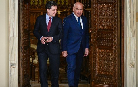 Nicușor Dan și Ilie Bolojan, ambii îmbrăcați în costume elegante, ieșind împreună dintr-o încăpere printr-o ușă masivă din lemn sculptat.