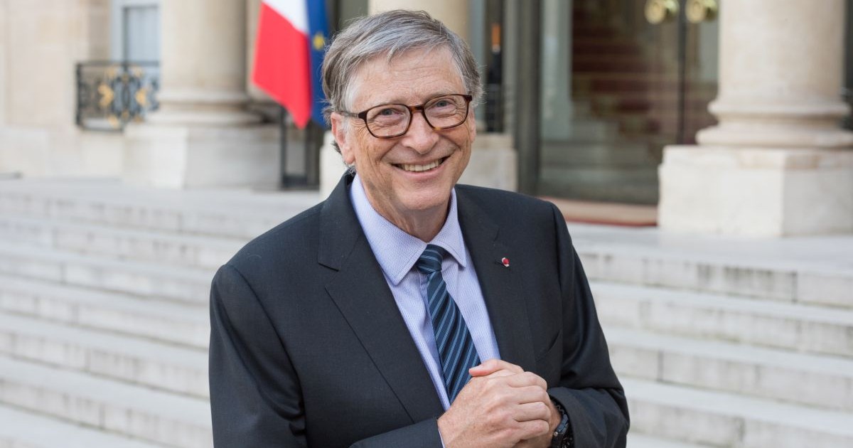 Bill Gates, in sacou inchis la culoare, camasa alba si cravata, tine maine impreunate in fata unei cladiri, in aer liber