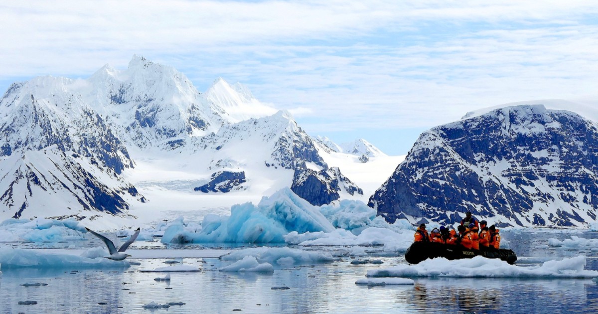 Un grup de turiști într-o barcă gonflabilă în Arctica, stau în barcă în timp ce ghidul le arată muntele de gheață din spatele lor, acesta este complet acoperit de zăpadă