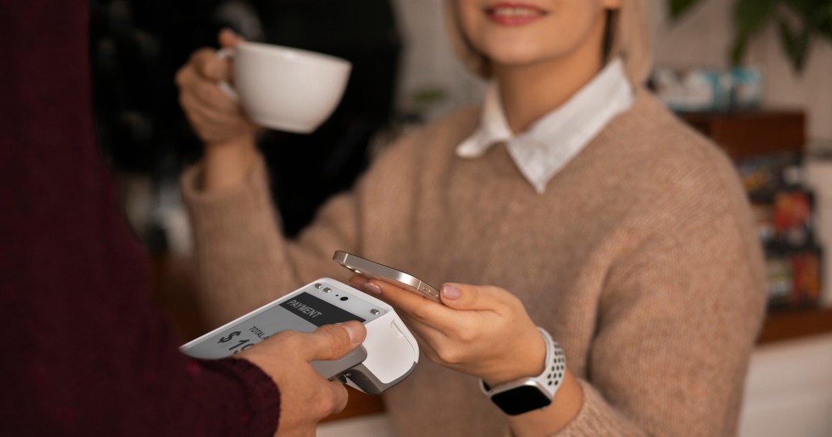 femeie îmbrăcată într-un pulover crem, cu guler alb de cămașă, ținând în mână o ceașcă albă de cafea și u card îndreptat spre un aparat POS în cealaltă mână