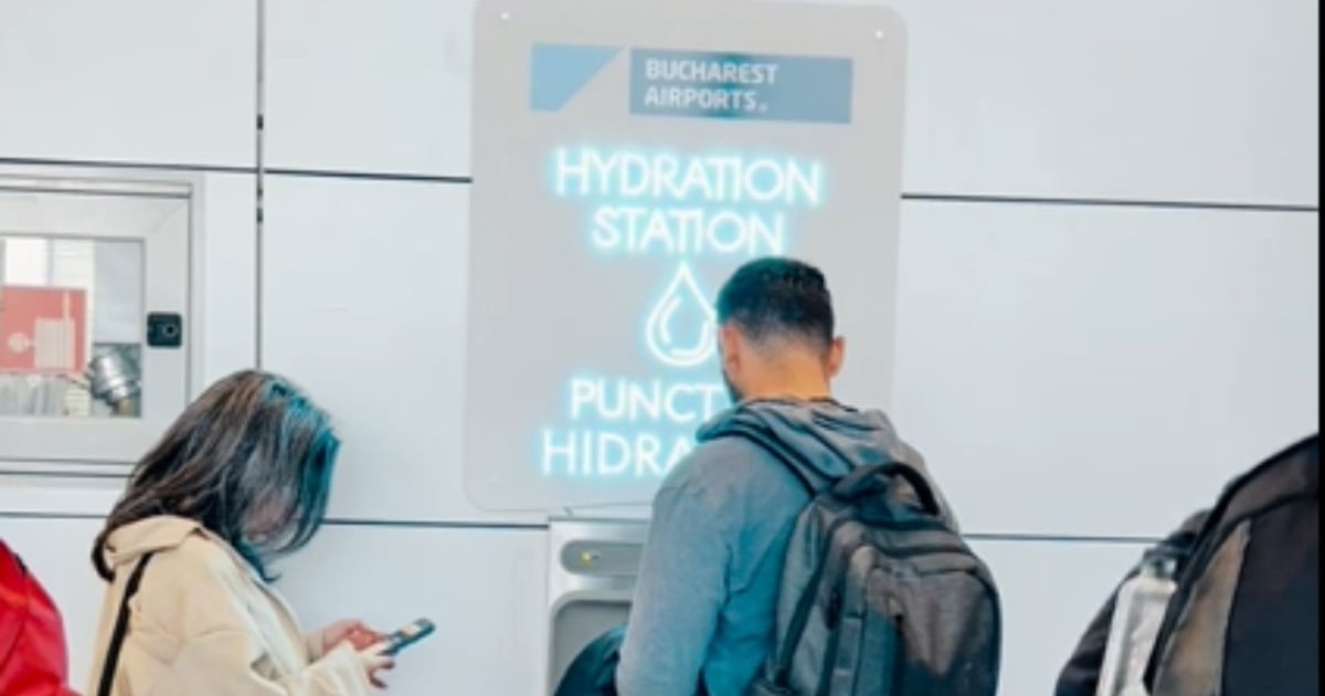 Imaginea surprinde un punct de hidratare instalat în interiorul unui aeroport, unde pasagerii își pot umple sticlele cu apă. În fața stației, mai multe persoane așteaptă sau își verifică telefoanele, în timp ce folosesc această facilitate.
