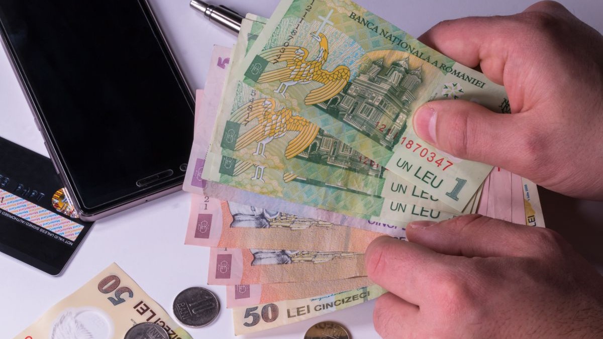 Un bărbat ține în mână mai mulți bani hârtii, iar pe masă mai sunt monede, un card bancar și un telefon.