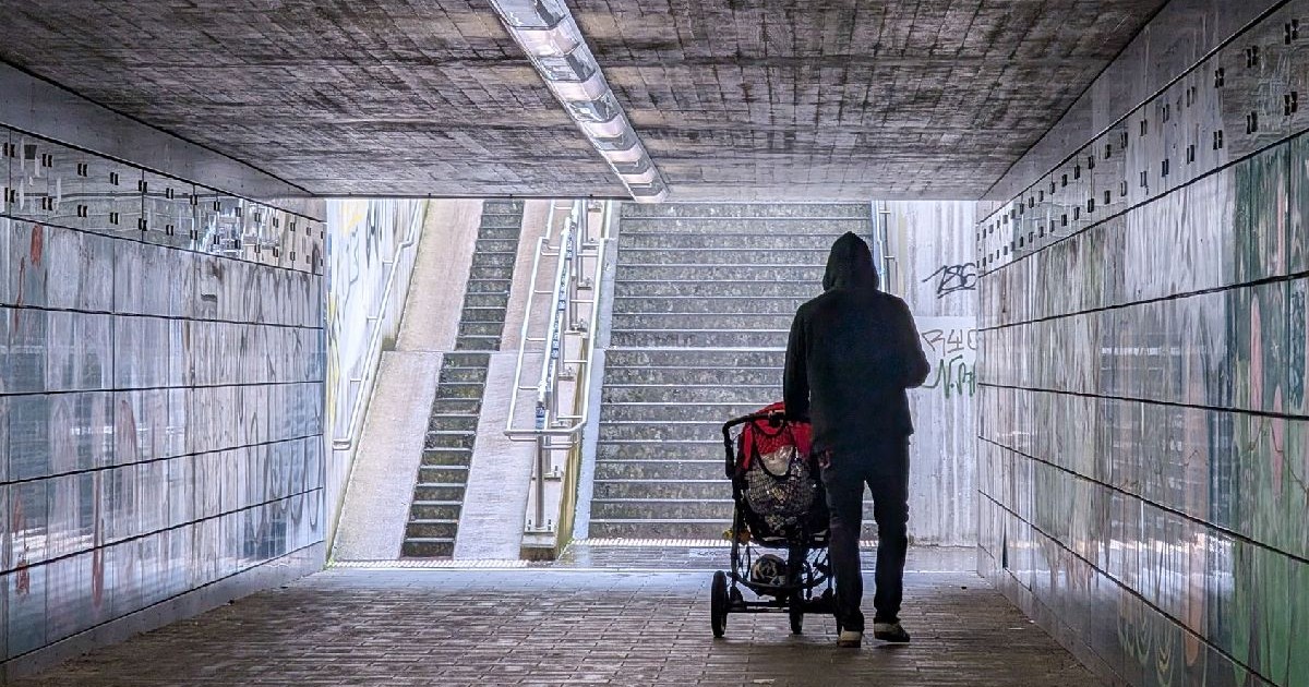 Imagine ilustrativă cu o persoană îmbrăcată într-un hanorac cu glugă împingând un cărucior printr-un tunel slab iluminat și acoperit cu graffiti, cu o scară în fundal.