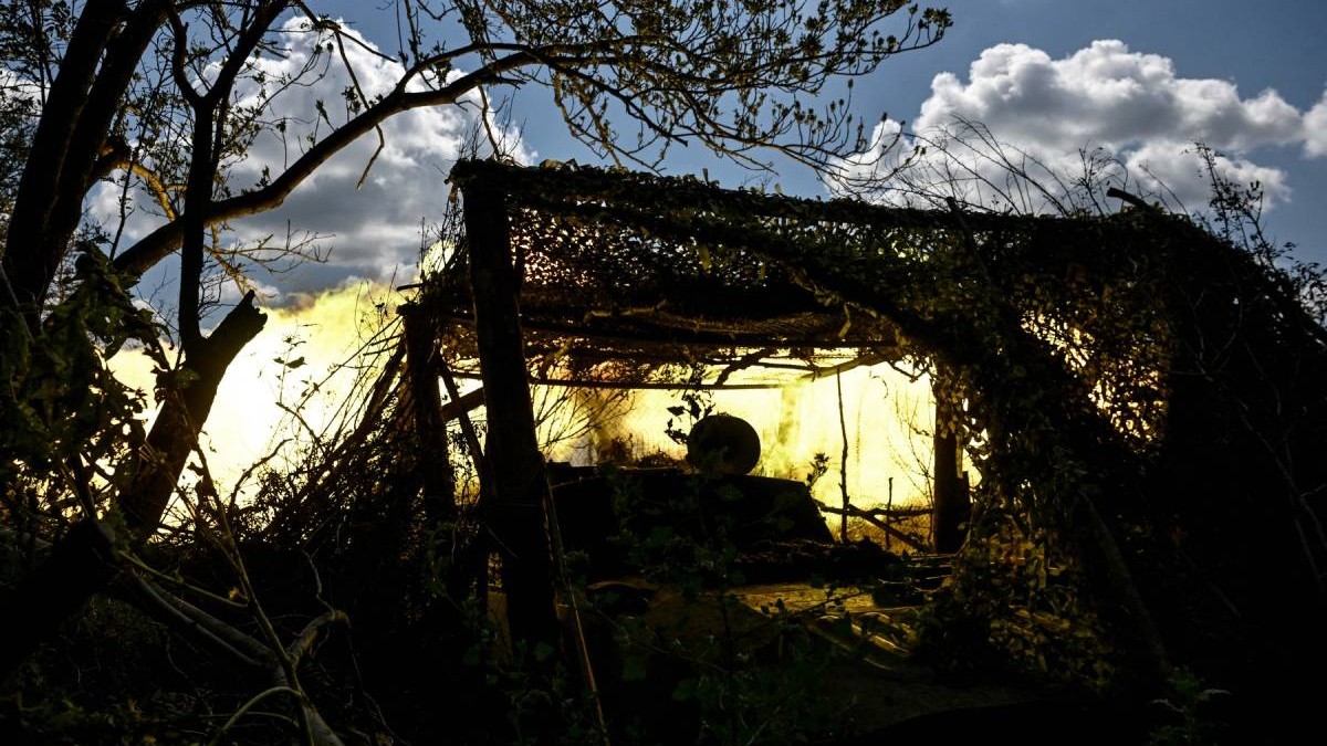 O poziție de artilerie camuflată în vegetație, luminată puternic de o explozie galben-aurie în fundal, cu crengi și frunziș care ascund silueta echipamentului militar în Pokrovsk - Ucraina